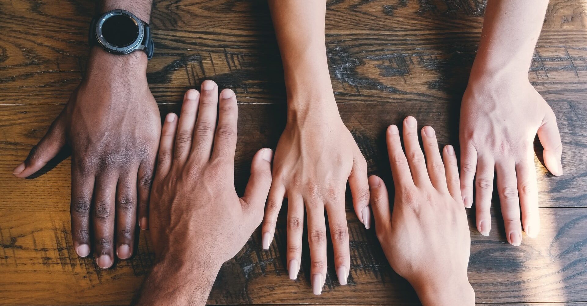 Five hands of diverse individuals placed side by side on a wooden table, symbolizing unity, teamwork, and diversity.