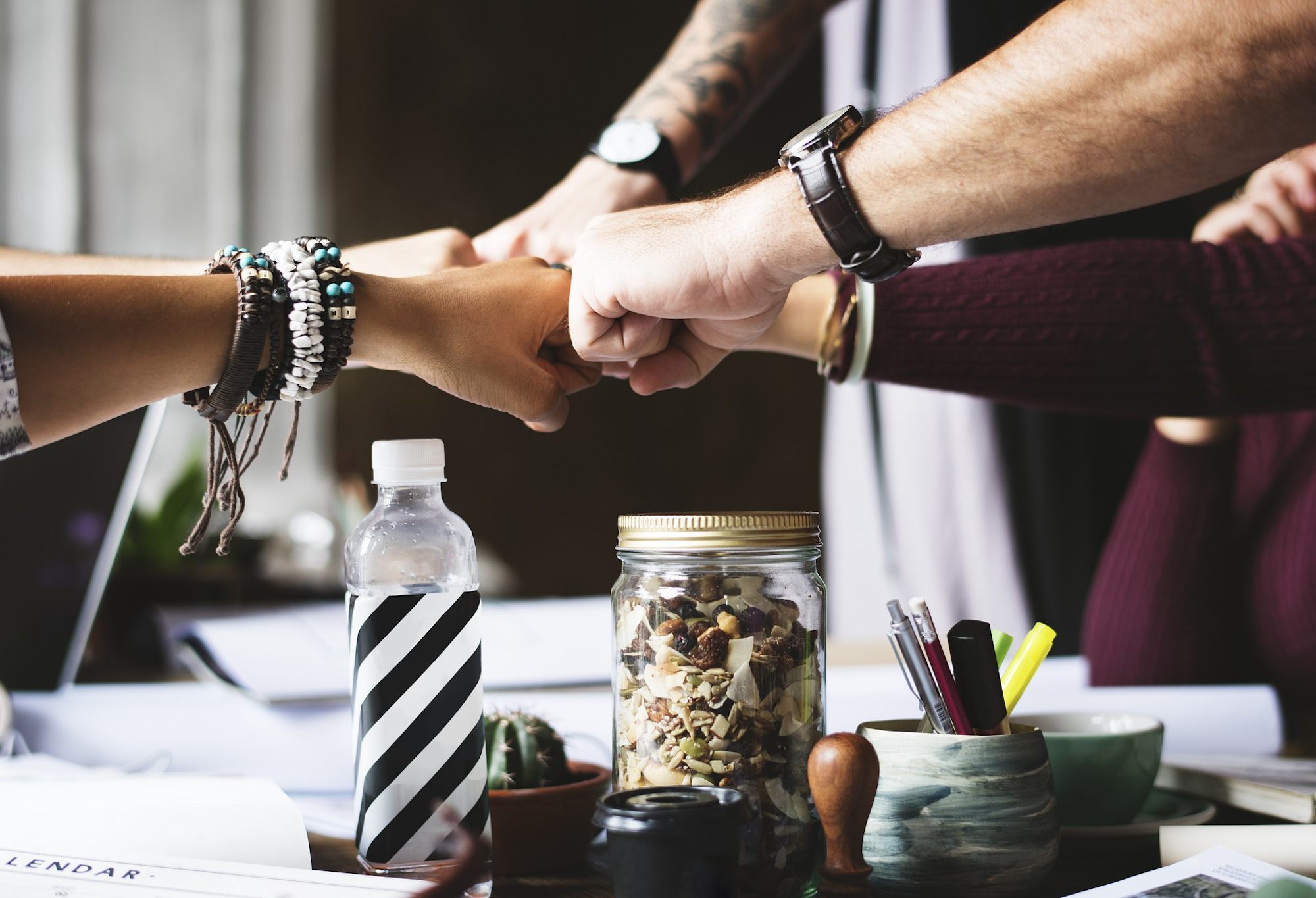 Group of people performing a fist bump over a table filled with office supplies, snacks, and drinks, symbolizing teamwork and unity in a collaborative workspace.