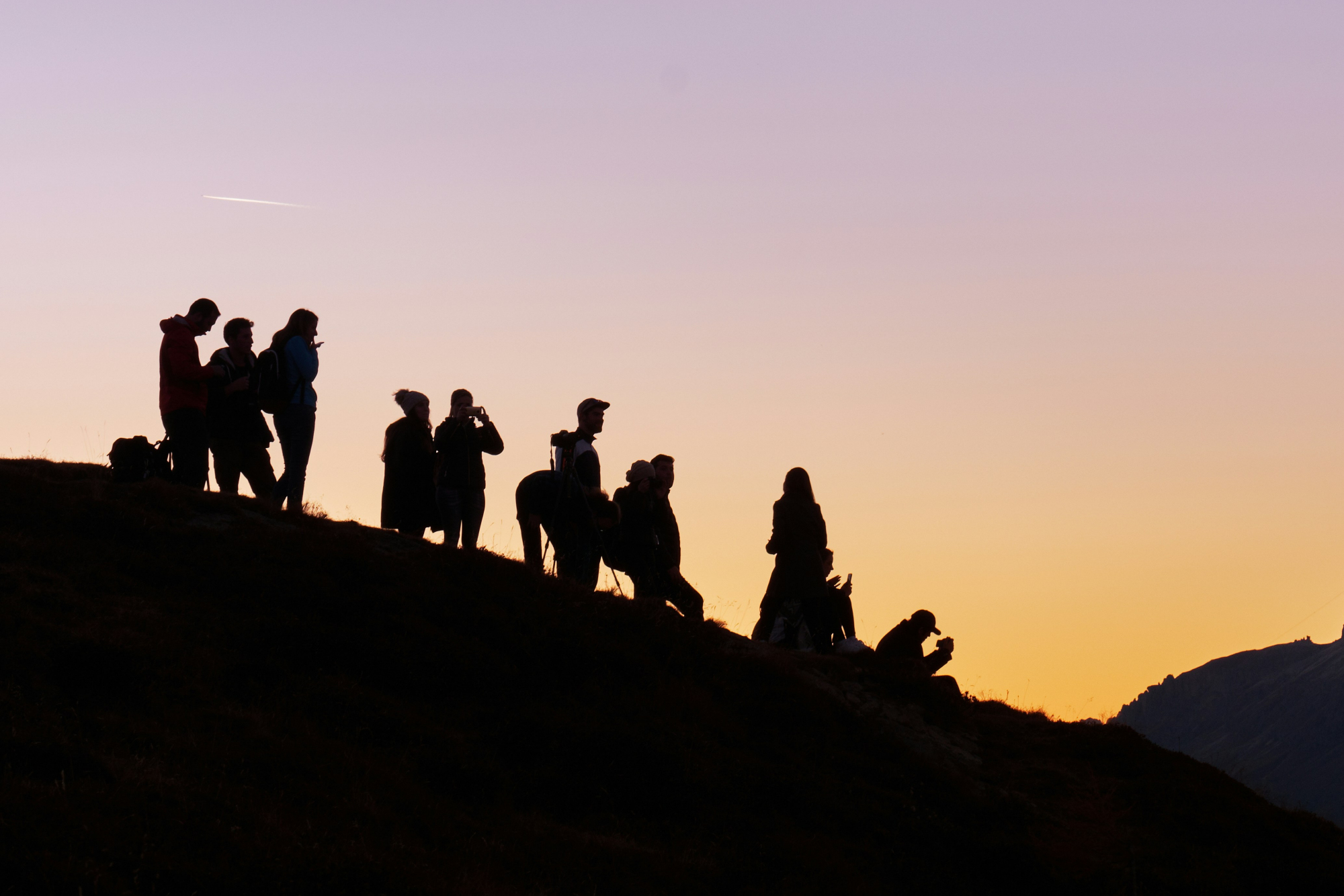 Silhouettes of a group of people standing on a hill at sunset, enjoying the view and capturing the moment, with a colorful sky in the background.