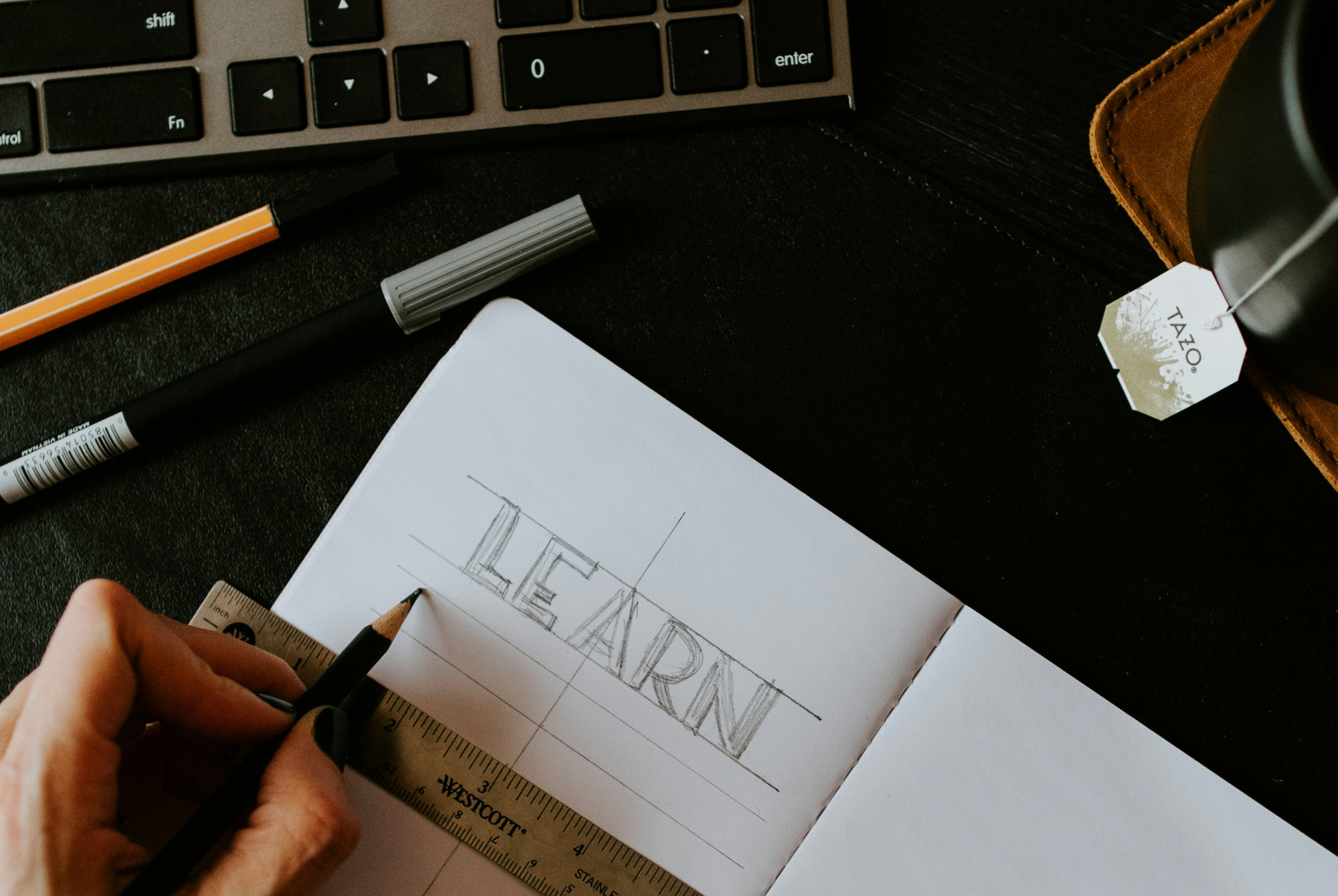 Person sketching the word "LEARN" in block letters on a notebook with a pencil and ruler, surrounded by a keyboard, pens, and a tea bag.