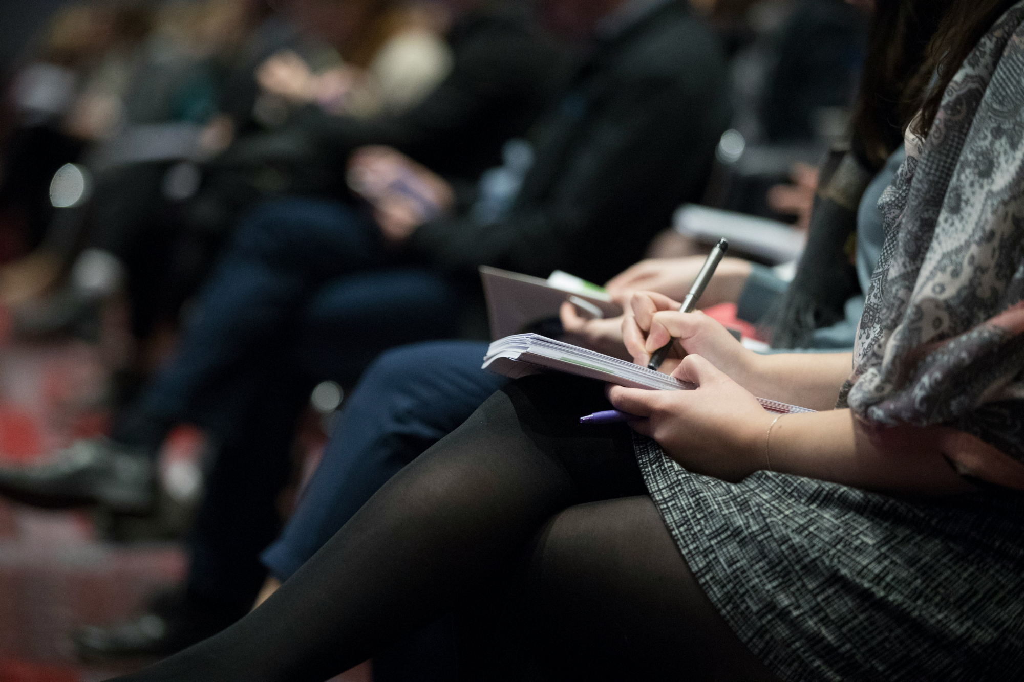 Audience members seated and taking notes during a conference or seminar, focusing on a close-up of a person writing in a notebook with a pen.