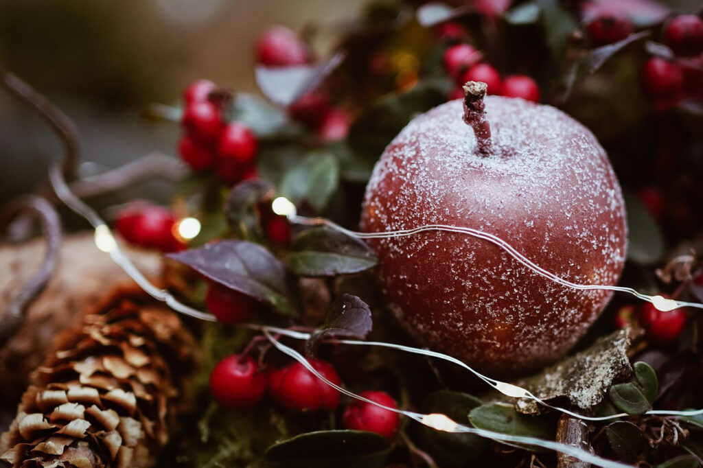 A Christmassy image, featuring a large frosted berry-shaped bauble, nested among green leaves, small red berries, and a thread of fairy lights