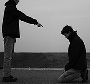 The culture challenge - a black and white photo of a bleak landscape with two people, one standing and pointing assertively at the bowed head of the other who is kneeling